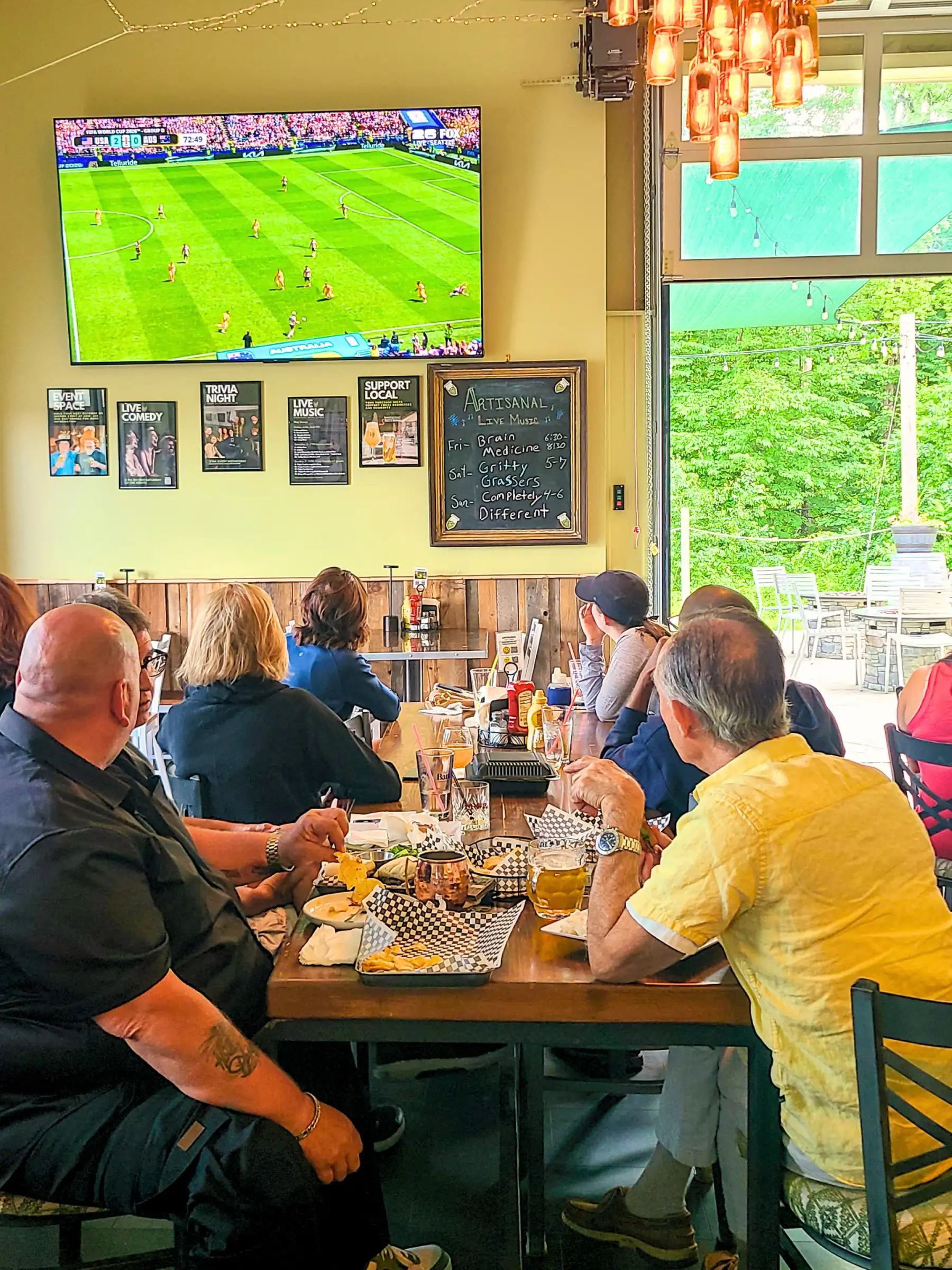 World Cup soccer viewing at Artisanal Brew Works in Saratoga Springs with guests watching a match on a 100-inch screen beside an open overhead door overlooking the forest.